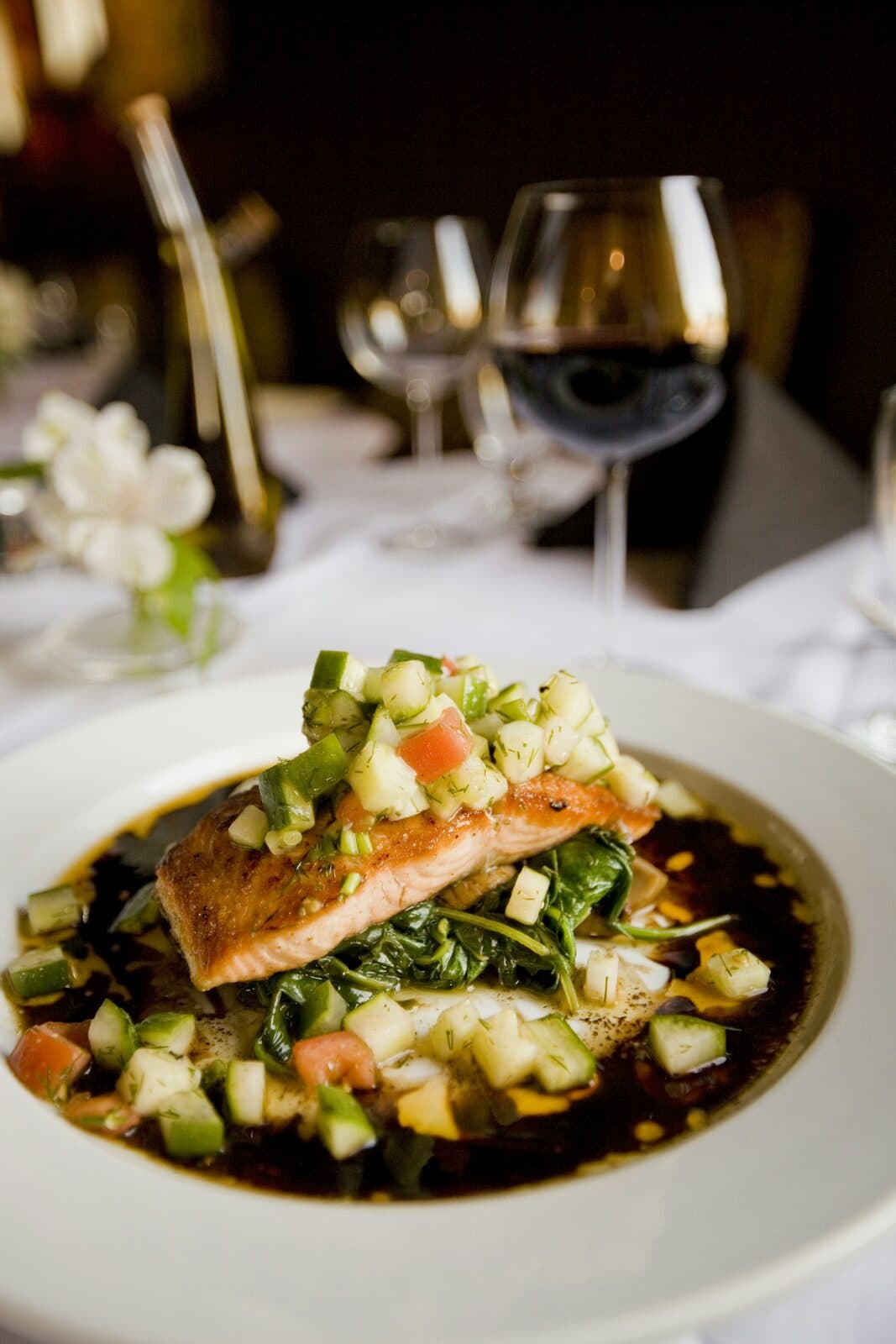 Plated salmon fillet on greens with a glass of red wine behind it in low light