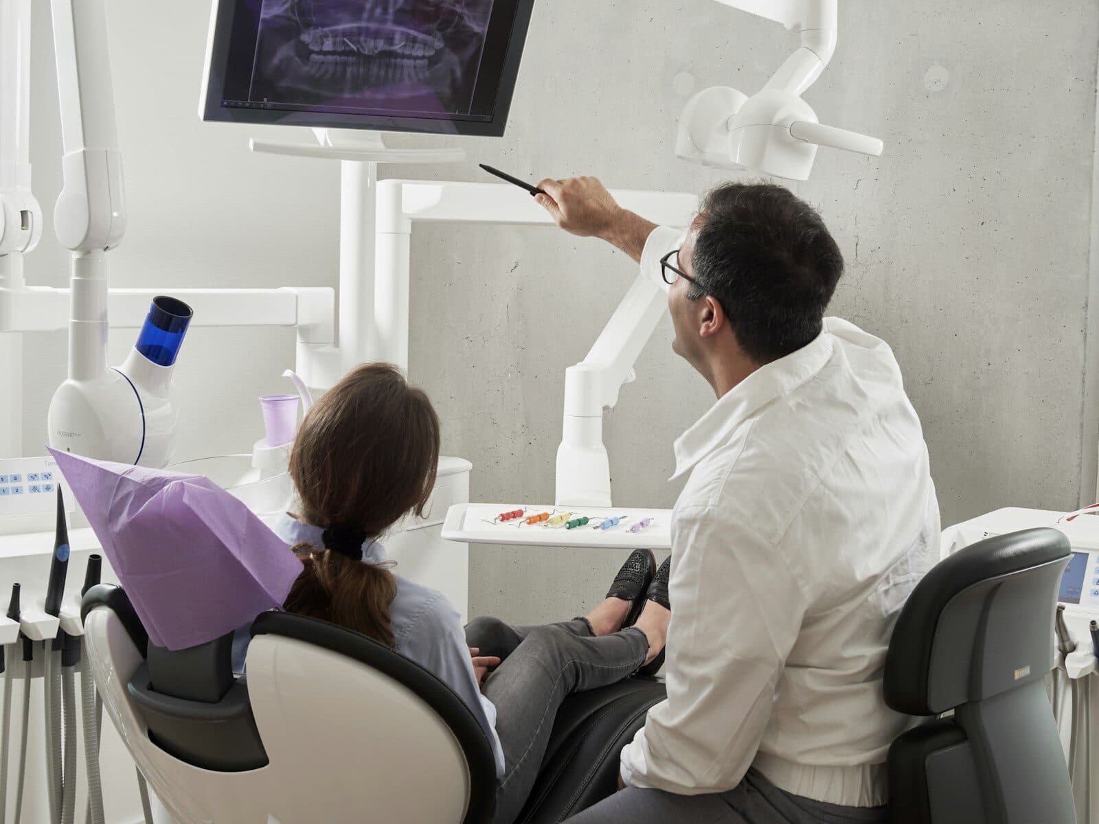 Dentist in a white coat pointing at a dental x-ray on a wall screen beside a patient in the chair