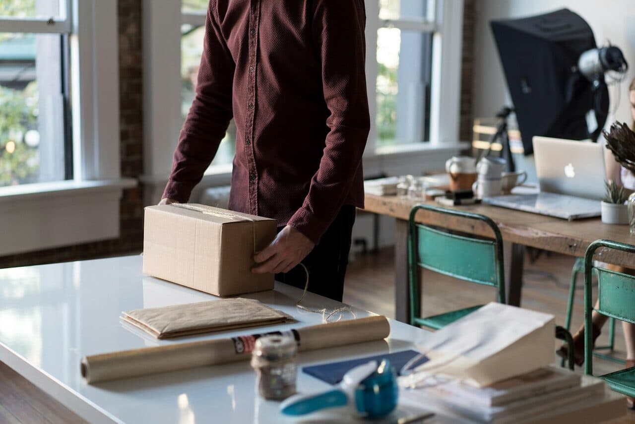 Person at a bright desk packing a small cardboard box beside a laptop